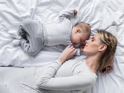 Top-down view of a mother wearing white pajamas sleeping peacefully next to her newborn baby on a white bed. This image accompanies a guide on navigating student loan repayment during maternity leave, illustrating how mothers can manage reduced income and rising household expenses through deferment, forbearance, or income-driven repayment strategies. Source: The College Investor