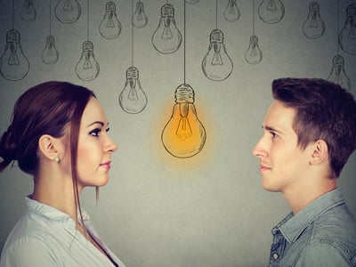 Profile views of a young woman and a young man facing each other, set against a grey background adorned with hand-drawn lightbulbs. A single bright yellow lightbulb glows centrally between them, symbolizing the realization or "bright idea" regarding the shifting gender dynamics in higher education. This visual metaphor accompanies an analysis of the growing gender gap in college degree attainment, where women now outpace men in earning Associate's, Bachelor's, Master's, and Doctoral degrees. Source: The College Investor
