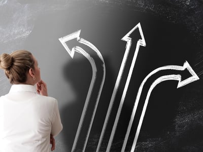 Rear view of a businesswoman in a white shirt looking up at a chalkboard with three large arrows pointing in different directions. This conceptual image illustrates the complex decision-making process parents face regarding student loans in 2026, as new federal limits on Parent PLUS loans force families to weigh federal versus private borrowing options. Source: The College Investor