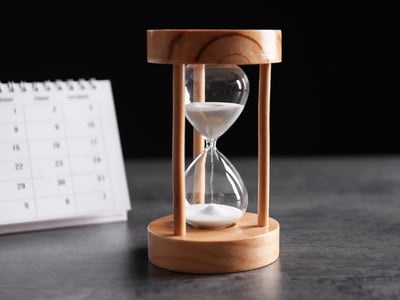 Close-up of a wooden hourglass with sand running out, sitting next to a white paper calendar on a dark surface. This image visualizes the urgency of the upcoming July 1, 2026, deadline for federal student loan borrowers—especially Parent PLUS holders—to consolidate their loans, or risk permanently losing access to income-driven repayment plans and forgiveness programs. Source: The College Investor