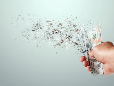 Close-up of a hand holding a stack of U.S. hundred-dollar bills that are disintegrating into dust and fragments on the left side. This striking visual metaphor illustrates the concept of money losing value or disappearing, reflecting the article's discussion on federal student loan subsidies and the government's projected losses per dollar lent under new reconciliation reforms. Source: The College Investor
