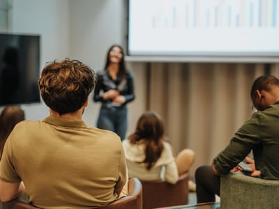 Rear view of a diverse group of adults sitting in a modern classroom, looking at a large screen displaying a colorful bar chart. A woman stands presenting, while attendees in casual attire focus on the data. This image illustrates the "workforce" aspect of the new Workforce Pell Grants, highlighting how adult learners can access federal funding for short-term, job-focused training programs in high-demand fields. Source: The College Investor