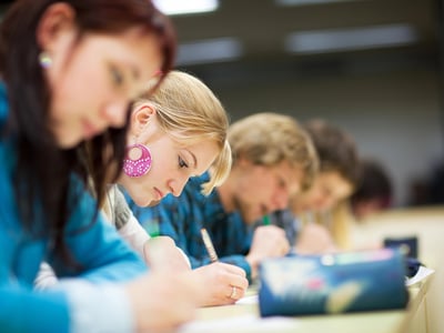 A row of focused high school students sitting at desks in a bright exam hall, diligently taking a written test. The camera's shallow depth of field highlights a young blonde woman in the center concentrating on her paper, while the students in front of and behind her are blurred. This scene of intense academic concentration illustrates the return of standardized testing, as major universities and Ivy League schools nationwide reinstate SAT and ACT score requirements for college admissions. Source: The College Investor
