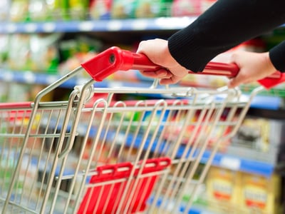 Close-up of a person's hands pushing an empty metal shopping cart with a red handle in a grocery store aisle. The image visualizes the concept of a "No-Buy" challenge, where participants deliberately avoid purchasing non-essential items to save money and break spending habits. Source: The College Investor