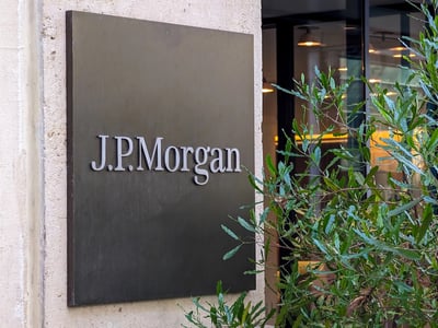 Close-up of a bronze corporate plaque reading "J.P.Morgan" mounted on a textured stone building facade next to a green leafy plant. This image accompanies a summary of J.P. Morgan Asset Management's 2026 College Planning Essentials report, which details the staggering 914% increase in college tuition since 1983 and the subsequent surge in student loan debt. Source: The College Investor
