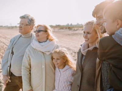 A multi-generational family of six, including grandparents, parents, and two young children, stands together on a sunny beach, smiling and looking out toward the horizon. They are dressed in warm, neutral-toned fall or winter coats and scarves. This image visually represents the concept of family support and interconnectedness, reflecting the article's focus on how rising student debt and housing costs are increasingly causing young adults to rely on their parents for financial help, impacting multiple generations' financial futures. Source: The College Investor