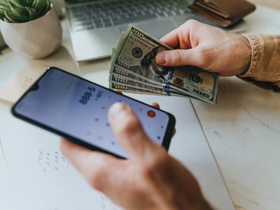 Close-up of a person's hands holding a smartphone displaying a calculator app in one hand and a stack of U.S. hundred-dollar bills in the other, set over a piece of paper with handwritten math. This image illustrates the financial calculation borrowers must make regarding the PSLF Buyback program, specifically highlighting how the recent court ruling blocking the REPAYE plan could result in higher out-of-pocket costs for those seeking student loan forgiveness. Source: The College Investor