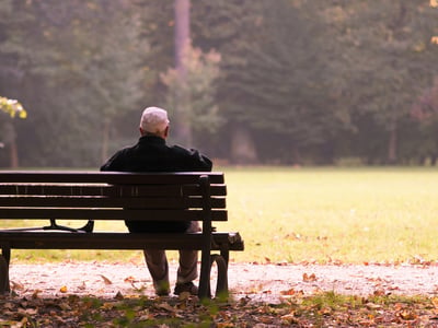 Rear view of an elderly man wearing a cap and jacket sitting alone on a park bench in autumn, with his cane resting beside him. The scene, framed by falling leaves and soft light, visually represents the solitude and financial uncertainty faced by retirees as traditional pension plans disappear across the healthcare and public sectors.