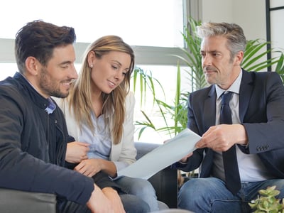 A smiling young couple sitting closely together on a grey sofa reviews financial documents handed to them by a professional advisor in a navy suit and blue tie. The three individuals are engaged in a positive discussion in a brightly lit, modern office setting with large windows and green plants. This image visualizes the consulting process for long-term financial planning, such as discussing early retirement strategies and setting up a Roth conversion ladder to access retirement funds without penalties. Source: The College Investor