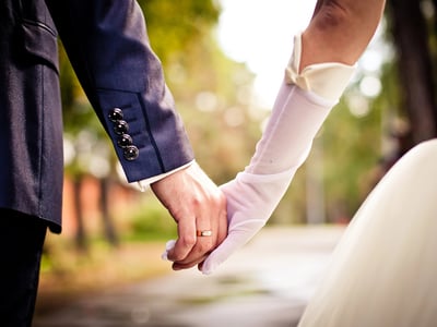 Close-up of a bride and groom holding hands, with the groom's hand displaying a simple gold wedding band. The bride is wearing a white dress and long sheer white gloves, while the groom wears a dark blue suit with button details on the cuff. The background is a soft, blurred outdoor setting with trees and sunlight. This image symbolizes the financial partnership of marriage, illustrating how couples can plan for the future together by utilizing tools like a spousal IRA to ensure a non-working spouse can still save for retirement. Source: The College Investor