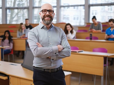 A smiling male college professor with a beard and glasses stands confidently with crossed arms in the foreground of a modern university lecture hall. Behind him, several students sit at wooden desks. This image represents the public service professionals—such as educators, non-profit workers, and government employees—who must navigate complex, changing federal repayment rules outlined in the PSLF Strategy Guide to successfully achieve Public Service Loan Forgiveness. Source: The College Investor