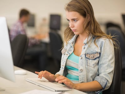 A focused female college student sits at a desktop computer in a campus lab, typing intently on a white keyboard. She wears a casual denim jacket over a striped shirt, representing a typical student balancing academics with extra work. In the blurred background, another person works at a separate workstation. This image illustrates the dedication and digital tools required to successfully launch and manage AI-powered side hustles to earn extra money during college. Source: The College Investor
