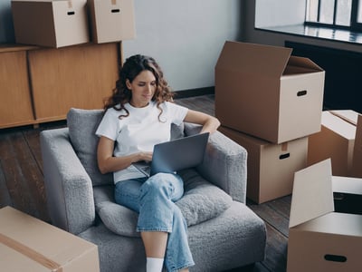 College student girl among cardboard boxes is using laptop and smiling.