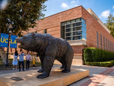 Man photographing bronze statue of Bruin with smartphone at campus of UCLA with college building in the background.