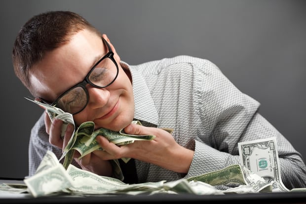 A man in a button-down shirt and glasses smiles contentedly while sleeping with his head resting on a large pile of U.S. dollar bills. This image illustrates the "sleep well at night" security of investing in 12-month Certificates of Deposit (CDs), which offer guaranteed returns with rates as high as 4.15% APY as of March 2026, allowing savers to rest easy knowing their money is growing safely. Source: The College Investor