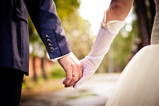 Spouse Close-up of a bride and groom holding hands, with the groom's hand displaying a simple gold wedding band. The bride is wearing a white dress and long sheer white gloves, while the groom wears a dark blue suit with button details on the cuff. The background is a soft, blurred outdoor setting with trees and sunlight. This image symbolizes the financial partnership of marriage, illustrating how couples can plan for the future together by utilizing tools like a spousal IRA to ensure a non-working spouse can still save for retirement. Source: The College Investor