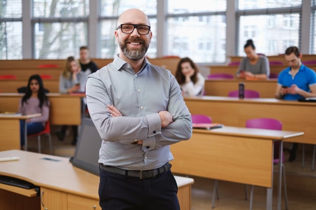 Teacher In a Classroom A smiling male college professor with a beard and glasses stands confidently with crossed arms in the foreground of a modern university lecture hall. Behind him, several students sit at wooden desks. This image represents the public service professionals—such as educators, non-profit workers, and government employees—who must navigate complex, changing federal repayment rules outlined in the PSLF Strategy Guide to successfully achieve Public Service Loan Forgiveness. Source: The College Investor