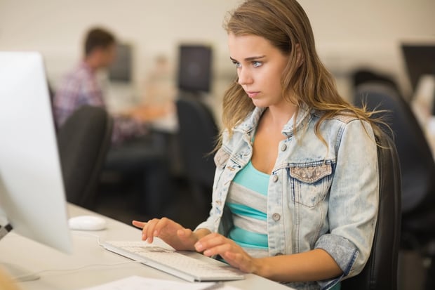 A focused female college student sits at a desktop computer in a campus lab, typing intently on a white keyboard. She wears a casual denim jacket over a striped shirt, representing a typical student balancing academics with extra work. In the blurred background, another person works at a separate workstation. This image illustrates the dedication and digital tools required to successfully launch and manage AI-powered side hustles to earn extra money during college. Source: The College Investor