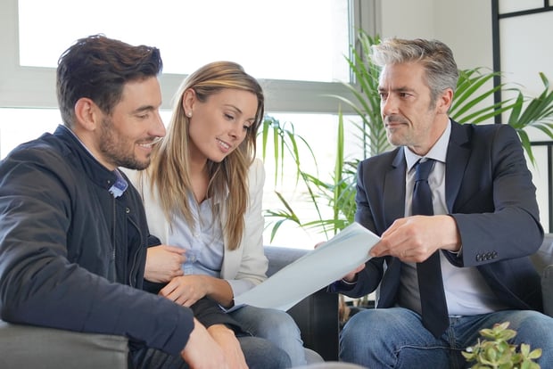 Couple looking over buyers agreement with real estate agent A smiling young couple sitting closely together on a grey sofa reviews financial documents handed to them by a professional advisor in a navy suit and blue tie. The three individuals are engaged in a positive discussion in a brightly lit, modern office setting with large windows and green plants. This image visualizes the consulting process for long-term financial planning, such as discussing early retirement strategies and setting up a Roth conversion ladder to access retirement funds without penalties. Source: The College Investor