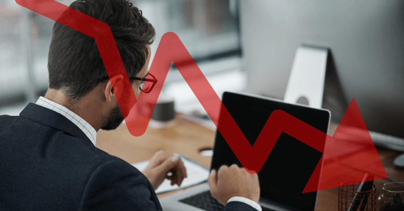 Declining Unemployment A dark-haired man in a suit and glasses is viewed from behind, intently looking at a laptop screen on a wooden desk in an office setting. The man's hands are resting near the keyboard, suggesting he is working or analyzing information. In the background, a modern desktop computer monitor is partially visible. Superimposed over the image is a prominent, thick, bright red arrow sharply declining from the upper left to the lower right corner, symbolizing a decrease or drop. This visual metaphor directly relates to the article's discussion of declining unemployment rates and the link between higher education and lower joblessness, illustrating the positive trend in the labor market despite changes to unemployment deferments and the need for new strategies in case of job loss.