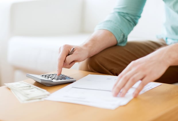 close up of man counting money and making notes A person in a light blue shirt and khaki pants sits at a light wooden table, actively calculating figures with a silver and black calculator. Their right hand points a finger at the calculator keys, while their left hand rests on a stack of white papers, possibly bills or financial documents. To the left of the calculator, a small stack of US dollar bills, likely twenty-dollar denominations, is visible, suggesting money management or financial planning. The background is softly blurred but shows a white sofa, indicating a home or office setting. This image visually represents the process of managing finances and calculating costs, directly relating to the article's focus on understanding and comparing student loan rates to save borrowers money over time. Source: The College Investor