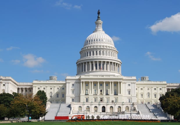 An eye-level, slightly upward-angled, daytime shot of the U.S. Capitol Building in Washington D.C. under a clear blue sky with sparse white clouds. The iconic white dome, topped with the Statue of Freedom, dominates the center of the frame, surrounded by its intricate columns and architectural details. The building's white marble facade extends horizontally, flanked by lush green trees on both sides, with more distant buildings partially visible. In the foreground, scaffolding and construction equipment are visible on the steps leading up to the Capitol, suggesting ongoing work. This image visually represents the "Congress" mentioned in the article, specifically the legislative body responsible for the "Big Beautiful Bill" that eliminates federal Grad PLUS loans, impacting graduate students' ability to afford professional programs like law and medicine.