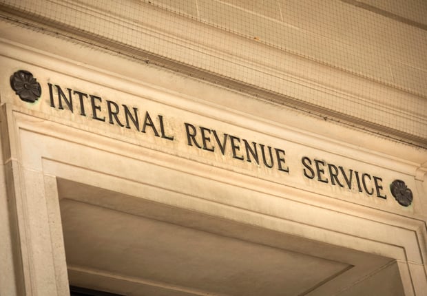 A close-up, slightly upward-angled shot of the Internal Revenue Service (IRS) federal building's entrance in Washington D.C., USA, emphasizing its prominent stone facade and the bold, black carved letters spelling "INTERNAL REVENUE SERVICE" above the doorway. The classic, ornate architectural details, including decorative dark rosettes flanking the text and intricately carved stone trim, are clearly visible. The image conveys a sense of official authority and directly relates to the article discussing the IRS's annual inflation adjustments for retirement accounts, specifically the raised 2026 contribution limits for 401(k) and IRA plans, as detailed in Notice 2025-67.