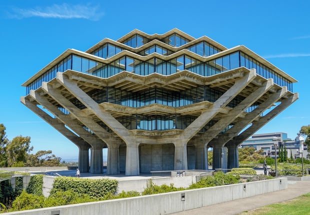 The iconic Geisel Library at the University of California San Diego stands majestically against a bright blue sky, its unique brutalist architecture resembling hands holding up a stack of books. This striking eight-story concrete and glass structure, with its geometric patterns and expansive windows reflecting the surroundings, is framed by lush green landscaping and pathways, hinting at the vibrant campus environment. The image visually anchors the article discussing concerns about UCSD freshmen's declining math skills and the university's response with remedial courses, raising questions about admissions practices, test-optional policies, and grade inflation amidst record demand for college admissions.