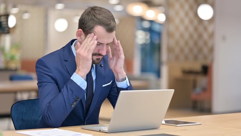 A visibly stressed middle-aged businessman, dressed in a sharp navy blue suit and tie, sits at a light wooden desk in a modern office environment, rubbing his temples with both hands as if experiencing a severe headache or intense frustration. His gaze is directed downwards towards an open silver laptop in front of him, suggesting he is overwhelmed by the information or tasks displayed on the screen. To his right, a tablet also rests on the desk, while a document is visible on his left. The soft-focus background shows a contemporary office with blurred lights and furniture, emphasizing the man's distress. This image effectively illustrates the article's theme of "Stressed by delayed technical issues," specifically the delayed implementation of the One Big Beautiful Bill Act (OBBBA) and its impact on Income-Based Repayment (IBR) for federal student loan borrowers. The visual conveys the anxiety and hardship caused by the postponement of crucial financial aid rule changes.