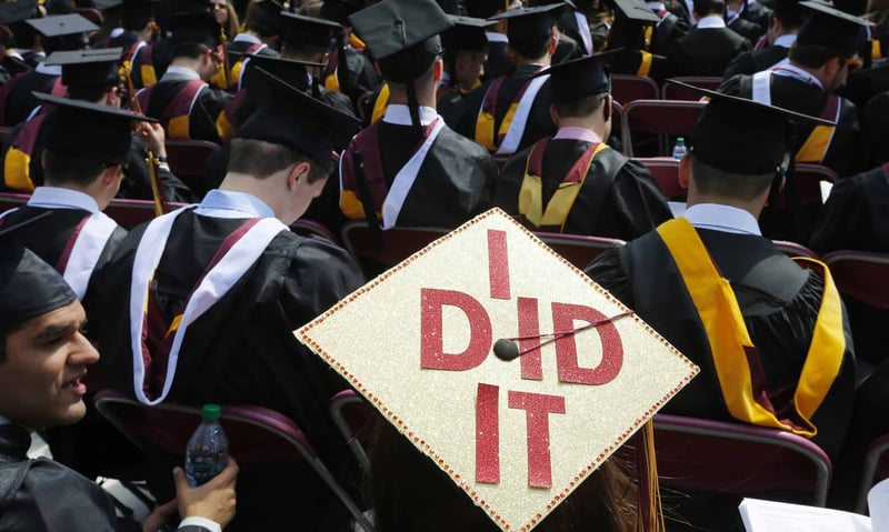 Rear view of a crowd of college graduates in black gowns and maroon-and-gold hoods sitting in rows during a commencement ceremony. In the center focus, a student wears a decorated mortarboard covered in gold glitter with large red sparkling letters spelling "I DiD IT." This celebratory scene marks the transition from student to borrower, the stage at which alumni begin repaying qualified student loans and may become eligible to claim the student loan interest deduction to reduce their taxable income. Source: The College Investor