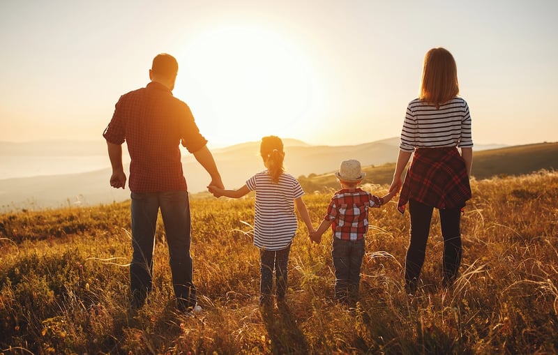 A family of four, including two adults and two young children, stands hand-in-hand facing away from the viewer, looking out over a sun-drenched, golden field at sunset or sunrise. The father, on the left, wears a plaid shirt and jeans, holding his daughter's hand. The daughter, in a striped shirt, holds her younger brother's hand, who is wearing a plaid shirt and a straw hat. The mother, on the right, wears a striped shirt, dark pants, and a plaid shirt tied around her waist, holding her son's hand. The warm, golden light of the sun, positioned centrally in the background, casts long shadows and illuminates the family and the expansive landscape of rolling hills and mountains in the distance. This image visually represents the "Family of Four Middle Class Squeeze" discussed in the article, highlighting the challenges middle-class families face with rising living costs and benefit cliffs, despite earning $100,000 to $200,000, as they struggle to get ahead financially. Source: The College Investor
