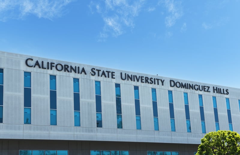 CARSON, CALIFORNIA - California State University Dominguez Hill sign on building. A wide, eye-level shot on a bright, clear day shows the modern, light-gray building of California State University Dominguez Hills under a blue sky with scattered wispy clouds. The university's name, "CALIFORNIA STATE UNIVERSITY DOMINGUEZ HILLS," is prominently displayed in large, dark blue, sans-serif letters across the top facade of the building. Below the signage, multiple rows of tall, rectangular windows with dark frames and reflective blue glass are evenly spaced, creating a pattern across the building's surface. In the bottom right corner, the vibrant green foliage of a tree is partially visible, adding a touch of nature to the academic setting. This image visually represents one of the California State University (CSU) campuses, providing context for the article's discussion of the new California law, Senate Bill 640 (SB 640), which will implement the Direct Admission Program for qualified California high school seniors starting in 2027. The law aims to boost college enrollment and ease the application process for students.