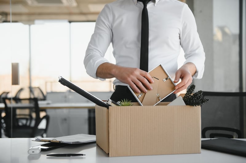 A close-up shot of a businessperson, dressed in a white shirt and black tie, carefully placing personal items into a cardboard box on a white office desk. The person's face is not visible, emphasizing the act of packing. Inside the box, a picture frame, a small potted plant, and decorative black berries are discernible, suggesting a recent termination or "tech layoff" as discussed in the article. The background shows a modern office environment with large windows, blurred office chairs, and desks, indicating a corporate setting. This image powerfully illustrates the current struggles faced by young workers in the tech industry, including college graduates and soon-to-be graduates, who are experiencing fewer job opportunities and a tougher path into the sector due to mass layoffs and a reshaping of early-career hiring patterns. Source: The College Investor