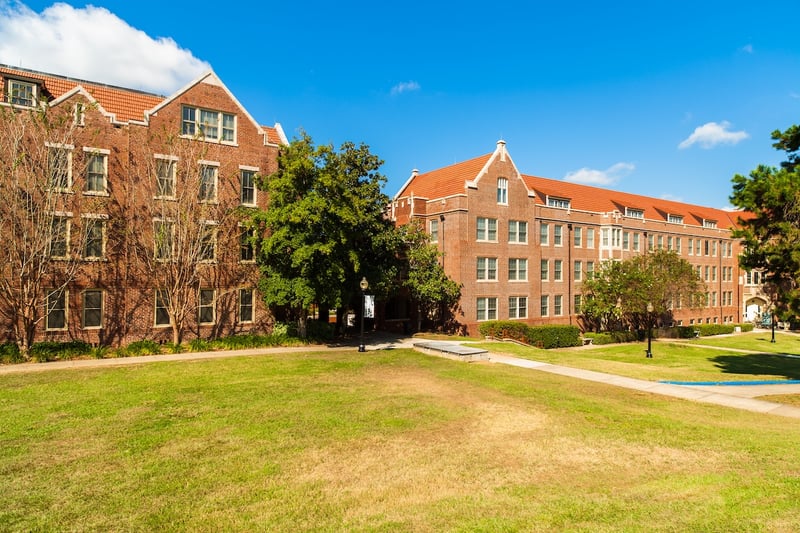 A bright, sunny view captures two imposing traditional red-brick college dormitory buildings with distinctive clay-tiled roofs, set against a vibrant blue sky with scattered white clouds. The multi-story dormitories feature numerous windows and are nestled within a well-maintained campus landscape, complete with manicured green lawns that sweep across the foreground and extend between the buildings. Several mature deciduous trees with lush green foliage are interspersed throughout the scene, casting soft shadows. A paved pathway winds through the grass, leading towards the dorm entrances. This visual representation highlights typical on-campus housing facilities, emphasizing the "room and board" expense which is a significant, and often overlooked, financial component of college costs that can even exceed tuition fees at many public universities. Source: The College Investor