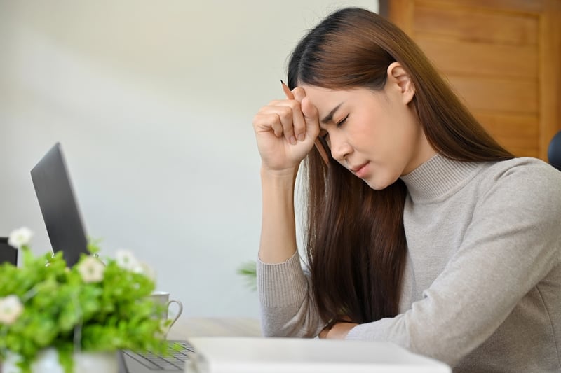 A side view of a young Asian woman with long brown hair, visibly stressed and overwhelmed, sitting at a light wooden desk. Her eyes are closed, her brow furrowed, and her right hand is pressed against her forehead, holding a dark pencil, indicating deep frustration or fatigue. A black laptop is open on the left side of the desk, along with a white coffee mug and a small green plant with white flowers, suggesting a home or office study environment. The background is a blurred light wall and a wooden door. This image powerfully illustrates the immense academic pressure, financial hardship, and mental health struggles that contribute to the alarming college dropout rate, where nearly 40% of students leave without earning a degree due to affordability issues, academic difficulties, and personal responsibilities. Source: The College Investor