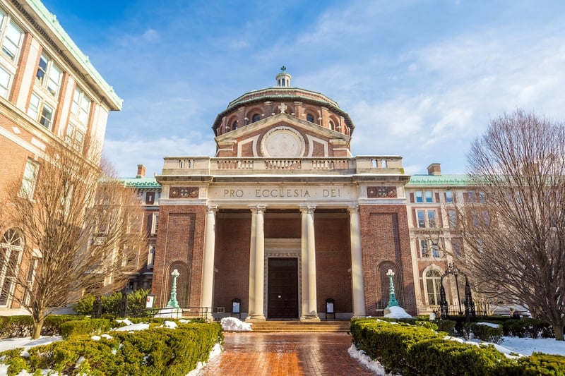 View of the red-brick and domed St. Paul's Chapel on the Columbia University campus, featuring a grand entrance with stone columns and the Latin inscription "Pro Ecclesia Dei." The snow-dusted Ivy League landmark represents the institution currently settling a class-action lawsuit for $9 million after allegedly submitting false data to inflate its U.S. News & World Report college ranking. Source: The College Investor