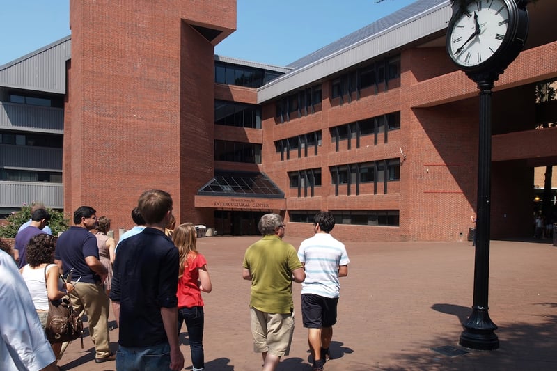 College Campus Tour at Georgetown University This image depicts a diverse group of prospective college students and their families participating in a campus tour at Georgetown University, an essential step in the college search process. The group is walking away from the viewer on a large, paved plaza towards a prominent brick building labeled "Intercultural Center," which is a central feature of the campus. The architectural style includes large windows and a multi-level design, with a modern clock on a tall black pole standing to the right. The bright sunny day suggests an ideal time for visiting schools and experiencing campus life firsthand, as discussed in the article regarding the importance of planning ahead and registering for tours to secure a spot and demonstrate interest to admissions officers.
