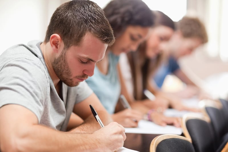 A focused group of diverse college students are diligently taking an important admissions exam, likely the Classic Learning Test (CLT), SAT, or ACT, as indicated by the article discussing these college entrance exams. The image shows several young adults seated at desks in a classroom setting, with the foreground focused on a male student with short brown hair and a beard, wearing a light grey t-shirt, intently writing on a paper with a black pen. Behind him, a female student with dark curly hair is also concentrating on her test, followed by other blurry figures of students suggesting a full classroom. This visual represents the academic rigor and the process high school students undergo when preparing for and taking these crucial standardized tests for college admission.