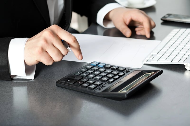 A close-up shot captures a person, likely a professional in a dark suit with a crisp white shirt, intensely focused on a black calculator with blue buttons while holding a pen. Their hands are positioned over the calculator and a blank white piece of paper, suggesting they are in the midst of financial calculations. To the right, a white computer keyboard and a smartphone are partially visible, indicating an office or study environment where important financial decisions are being made. This image visually represents the process of calculating the Public Service Loan Forgiveness (PSLF) Buyback amount, where borrowers determine what they owe to receive credit for past deferment or forbearance months, aligning with the article's focus on understanding and navigating the PSLF Buyback program.