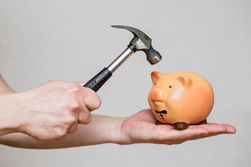 This close-up image features a person's outstretched hand holding a small, orange ceramic piggy bank, which is positioned directly below a silver hammer with a black handle. The hammer is held by the other hand, poised as if to strike the piggy bank, symbolizing the act of "cashing out" or breaking into savings. The piggy bank itself is detailed with small ears, a snout with two nostrils, and dark eyes, giving it a friendly appearance, contrasted by the implied action of the hammer. The background is a plain, light grey, ensuring full focus on the hands, hammer, and piggy bank. This visual metaphor directly relates to the article's discussion on how to cash out a life insurance policy, such as a Gerber Grow-Up Plan, and the implications of accessing its cash value for purposes like college costs, highlighting the decision to tap into accumulated funds.