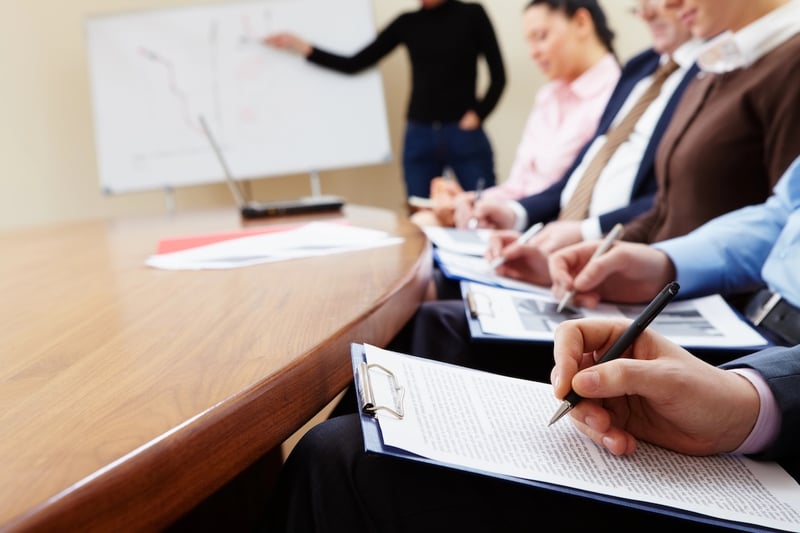 A blurry, close-up shot of a group of professionals attending a training or briefing session in an office conference room, underscoring the article's focus on Training Repayment Agreement Provisions (TRAPs). In the foreground, a person's hand is visible, holding a black pen and writing notes on a document attached to a clipboard, emphasizing the active learning and contractual nature of job training. Other attendees are partially visible in the middle ground, also taking notes, with one individual in a light pink shirt and another in a brown top. In the background, out of focus, a presenter in a black shirt gestures towards a whiteboard, which has diagrams or notes written on it, symbolizing the "training costs" discussed in the article. The warm wooden conference table dominates the left side of the frame, grounding the professional setting. This image visually represents the formal training environments where TRAPs are often implemented, highlighting the contracts and agreements workers sign that can lead to significant debt if they leave their employment early.