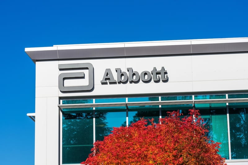 Abbott Laboratories This image features the prominent gray logo and wordmark "Abbott" on the clean, white facade of an Abbott Laboratories building, signifying the company's corporate identity. Below the logo, a striking tree with vibrant red and orange autumn foliage adds a natural contrast against the modern architecture and the clear, bright blue sky above. The building's windows reflect the sky and surrounding environment, adding depth to the scene. This visual serves to contextualize Abbott Laboratories as one of the major U.S. employers offering retirement plan matches tied to student loan payments, a key point discussed in the accompanying article about the SECURE 2.0 provision. Source: The College Investor