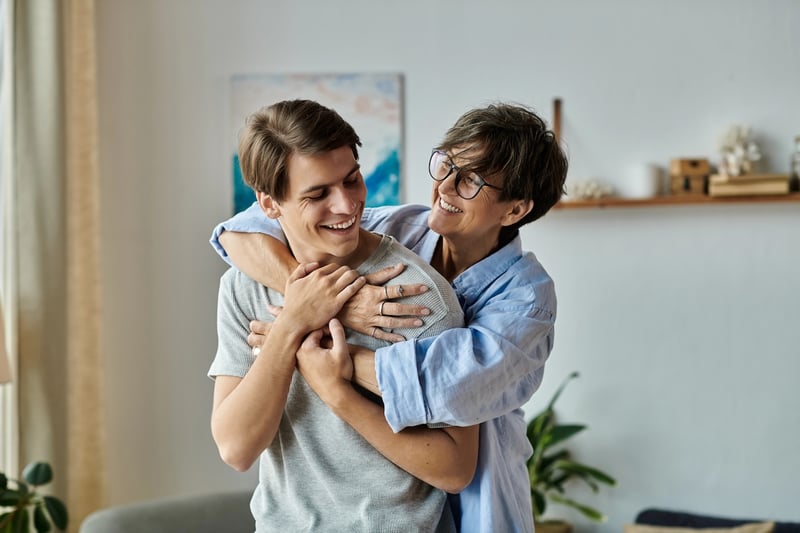 A loving mother with short, dark, salt-and-pepper hair and stylish round glasses, wearing a light blue button-down shirt, embraces her adult son from behind in a warm, brightly lit home. Both are smiling broadly, looking at each other affectionately, reflecting a close family relationship. The son, with short brown hair and a grey t-shirt, is facing forward, his hands resting gently on his mother's arms wrapped around him. This image illustrates the close bond between parents and adult children, which is central to the article discussing why shared bank accounts can create legal, tax, and financial aid risks as children become adults. The cozy home setting features blurred background elements like a light-colored wall, a painting with blue hues, and a plant, suggesting a comfortable, personal environment where financial discussions like those in the article might take place. Source: The College Investor