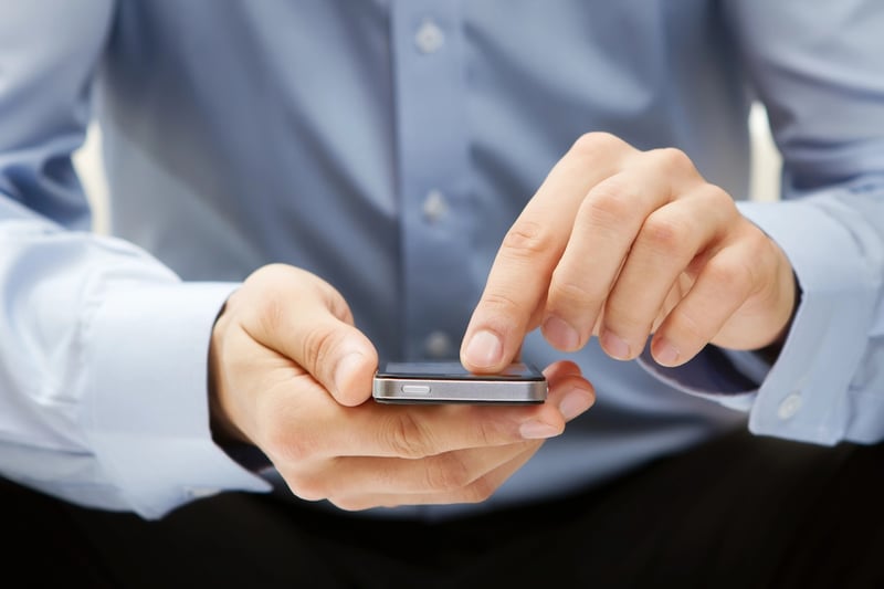 A close-up shot shows a person wearing a light blue dress shirt, with the sleeves rolled up, holding a silver smartphone horizontally with both hands. Their thumbs are positioned over the screen, poised to interact with the device, which glows faintly. This image directly relates to the article's discussion of the "vice economy," where mobile gambling and sports betting apps blur the lines between entertainment and finance for young adults. The phone represents the ease of access to these activities, contrasting with traditional financial advice and highlighting how social media, betting apps, and speculative investing contribute to a culture of instant gratification. Source: The College Investor