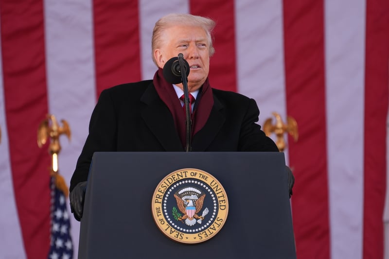 President Donald Trump, with his signature blonde hair, stands authoritatively at a dark podium adorned with the official Seal of the President of the United States. He is dressed in a smart black overcoat and a rich maroon scarf, speaking into a microphone positioned on the podium. Behind him, large American flags with prominent red and white stripes create a patriotic backdrop, flanked by golden eagle finials atop flagpoles. This image visually represents the executive branch's influence, illustrating the context of the article discussing the Trump administration's efforts to block the Consumer Financial Protection Bureau (CFPB) from accessing Federal Reserve funds, a move that could deplete the agency's budget by early 2026 and potentially lead to its effective closure. Source: The College Investor