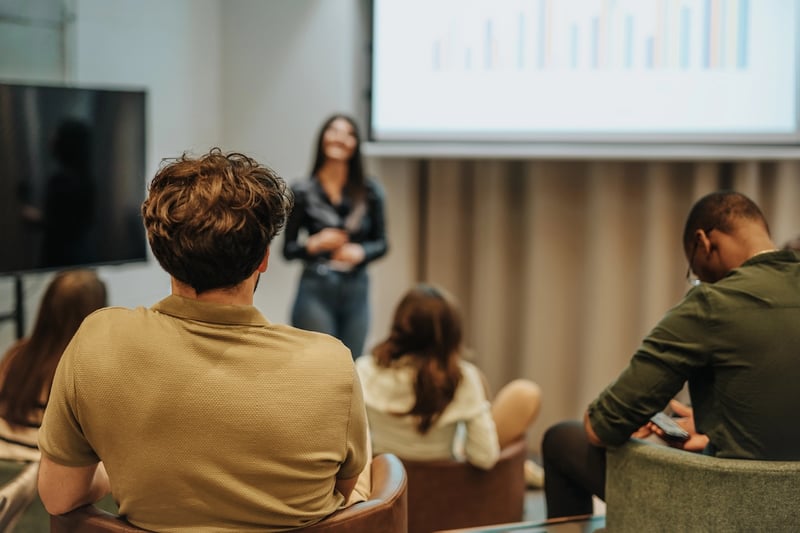 Rear view of a diverse group of adults sitting in a modern classroom, looking at a large screen displaying a colorful bar chart. A woman stands presenting, while attendees in casual attire focus on the data. This image illustrates the "workforce" aspect of the new Workforce Pell Grants, highlighting how adult learners can access federal funding for short-term, job-focused training programs in high-demand fields. Source: The College Investor