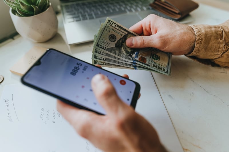 PSLF Buyback Math Close-up of a person's hands holding a smartphone displaying a calculator app in one hand and a stack of U.S. hundred-dollar bills in the other, set over a piece of paper with handwritten math. This image illustrates the financial calculation borrowers must make regarding the PSLF Buyback program, specifically highlighting how the recent court ruling blocking the REPAYE plan could result in higher out-of-pocket costs for those seeking student loan forgiveness. Source: The College Investor