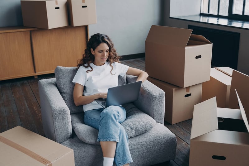 College student girl among cardboard boxes is using laptop and smiling.