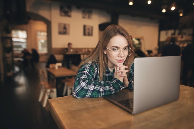 Beautiful brunette using notebook in cafe A young, light-skinned woman with long, straight brown hair and round, gold-rimmed glasses intently uses a silver laptop in a cozy, somewhat dimly lit cafe or co-working space. She is wearing a green and blue plaid shirt and rests her chin on her clasped hands, leaning forward slightly as she focuses on the screen. The background is blurred, showing the warm, inviting atmosphere of the cafe with wooden tables, chairs, and other patrons, suggesting a focused study or work session. This image illustrates a student utilizing modern tools like AI for scholarship searches, as discussed in the article, highlighting the blend of traditional study environments with advanced technology to streamline the process of finding scholarships and grants for college.