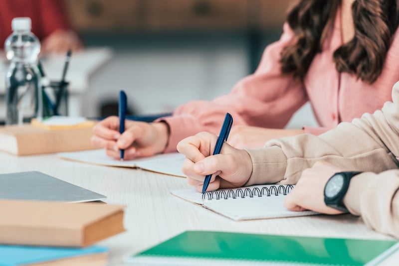 A close-up shot captures two individuals, likely students, diligently writing in notebooks with blue pens, illustrating the academic context relevant to the article's discussion of the Department of Education. The foreground shows two sets of hands; one hand, wearing a dark-faced wristwatch, writes in a spiral-bound notebook, while another hand, further back, writes in a different notebook. Both are using blue pens. The person in the background wears a long-sleeved pink top, while the person in the foreground wears a beige or light brown long-sleeved top. Books, a laptop, and a water bottle are visible on the light-colored desk, suggesting a study or learning environment. This image visually supports the article's focus on student loan forgiveness, Income-Driven Repayment (IDR), and Public Service Loan Forgiveness (PSLF) issues affected by the recent lawsuit settlement.